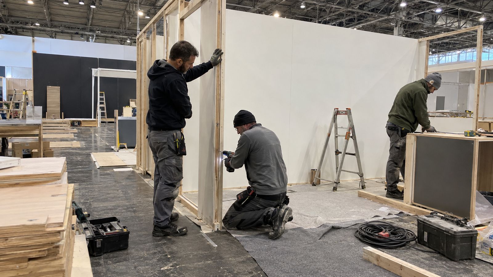 Stand installers assemble timber panels and a display counter inside an exhibition hall.
