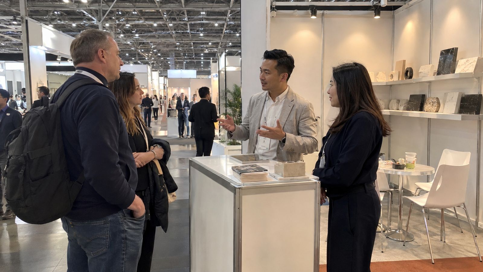 A host and interpreter welcome visitors and assist a conversation at an exhibition stand.