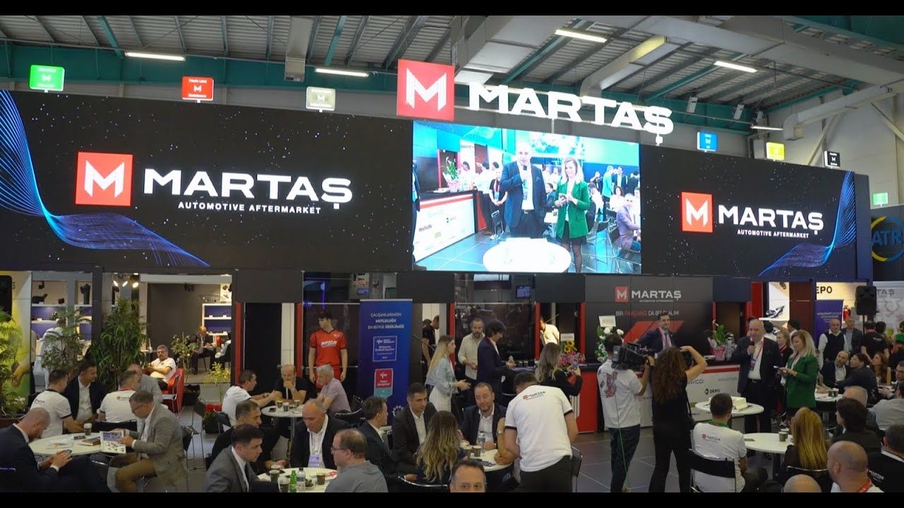 Visitors in discussions at tables in an exhibition stand beneath a large LED screen