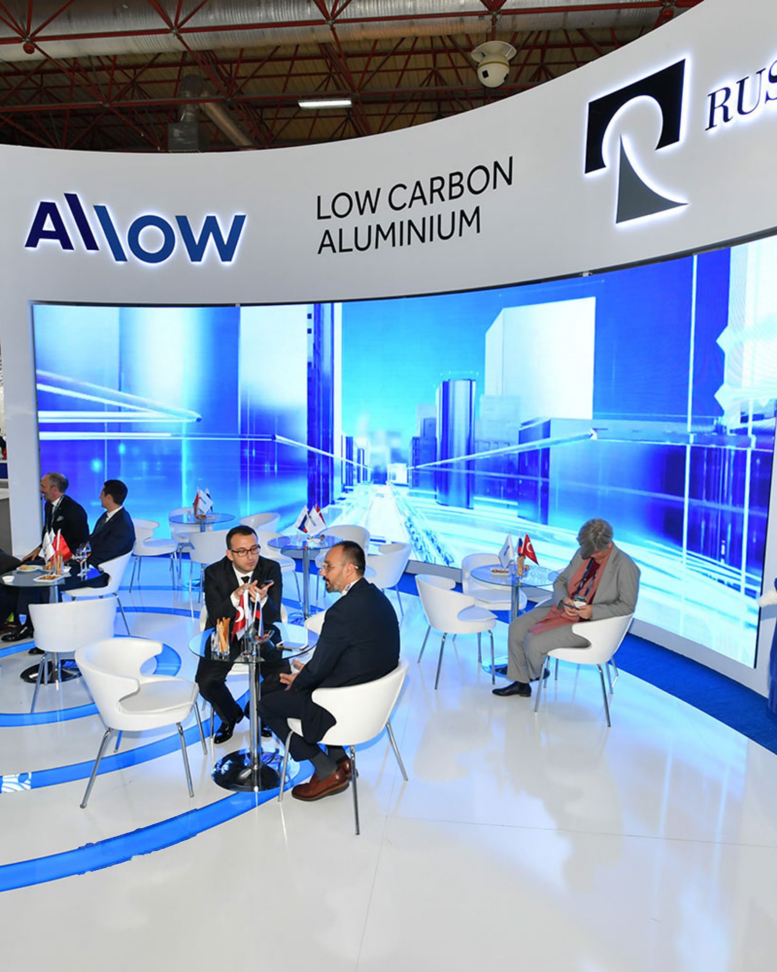 Visitors holding discussions at tables in a white and blue exhibition stand in front of a large screen