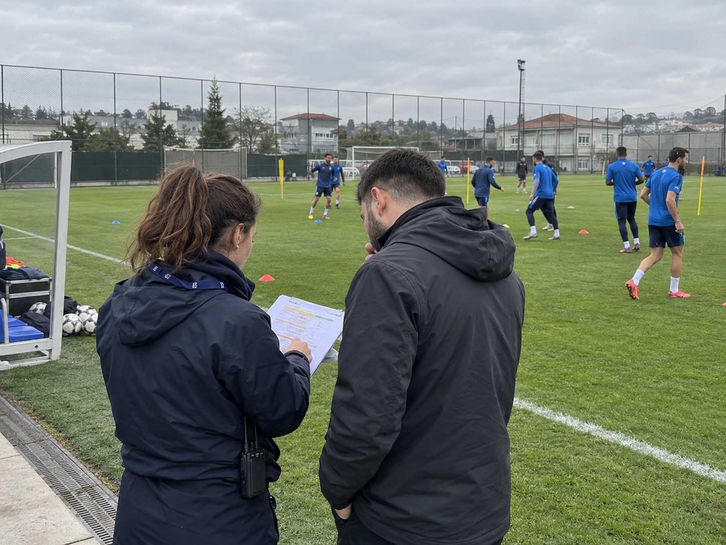 Two coordinators check a running order beside a pitch while football players warm up.